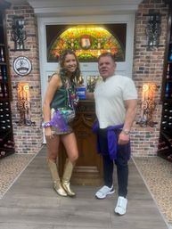 Two people smiling and posing inside a brick-walled wine room or tasting area with a colorful stained-glass window and wooden lectern behind them; the woman wears a metallic skirt, gold cowboy boots and layered purple, green and gold Mardi Gras beads, the man wears a white shirt, dark pants, white sneakers and a purple sweatshirt tied at his waist.