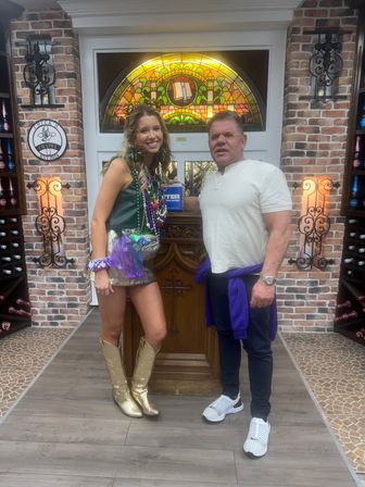 Two people smiling and posing inside a brick-walled wine room or tasting area with a colorful stained-glass window and wooden lectern behind them; the woman wears a metallic skirt, gold cowboy boots and layered purple, green and gold Mardi Gras beads, the man wears a white shirt, dark pants, white sneakers and a purple sweatshirt tied at his waist.
