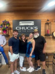 Group of four smiling adults posing inside a wine-and-spirits shop interior with a large black wall sign on wood paneling, colorful Mardi Gras bead wreath, lantern decor and bar stools.