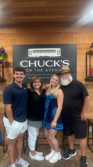 Group of four smiling adults posing inside a wine-and-spirits shop interior with a large black wall sign on wood paneling, colorful Mardi Gras bead wreath, lantern decor and bar stools.