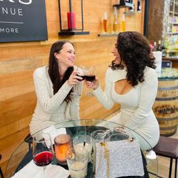 Two friends clinking red wine glasses at a cozy wooden-walled wine bar, laughing across a glass table with candles, cocktails and a sparkly handbag — wine tasting, girls’ night out vibe.