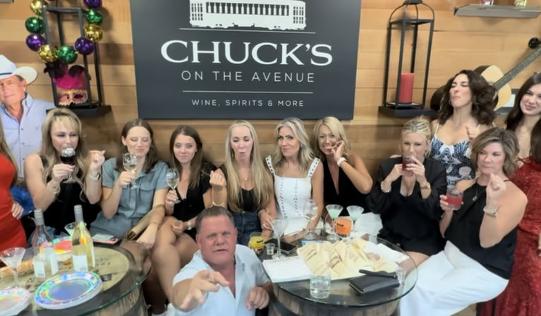 Group at an indoor wine-tasting event in a wood-paneled tasting room: a man reaches toward the camera in front while a row of women behind hold wine glasses and cocktails around barrel tables with bottles and snacks.