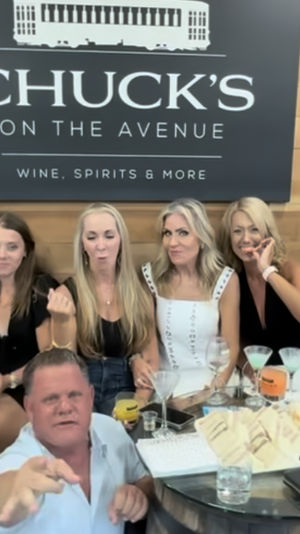 Group at an indoor wine-tasting event in a wood-paneled tasting room: a man reaches toward the camera in front while a row of women behind hold wine glasses and cocktails around barrel tables with bottles and snacks.