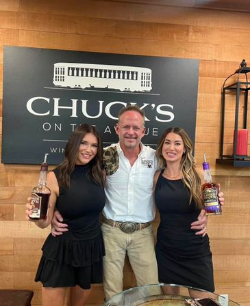 Three smiling people (two women and one man) posing in a wood-paneled whiskey tasting room, each holding a bottle of spirits in front of a large black wall sign — friendly group photo.