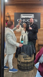 Three people raising champagne flutes in a celebratory toast at a rustic indoor tasting room, glass-top barrel table with bottle and wood-paneled wall backdrop.