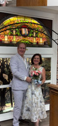 Smiling wedding couple indoors beneath a colorful arched stained-glass window; bride in floral embroidered dress holding a pink bouquet, groom in a light seersucker suit.