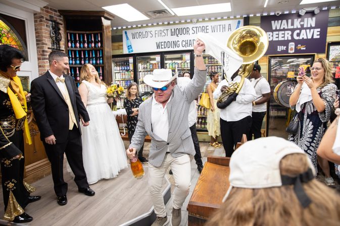 Lively wedding celebration inside a liquor store with the bride and groom smiling as a man in a hat dances holding a beer bottle, a brass band playing, and guests cheering near store coolers and a walk-in beer cave sign.