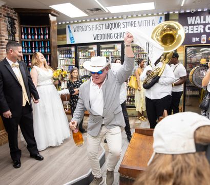 Lively wedding celebration inside a liquor store with the bride and groom smiling as a man in a hat dances holding a beer bottle, a brass band playing, and guests cheering near store coolers and a walk-in beer cave sign.