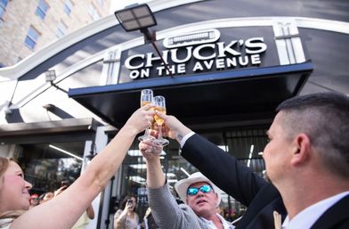 Four people clinking champagne flutes in a celebratory toast outside a black-and-white restaurant storefront on a busy city avenue, with onlookers and urban buildings in the background.