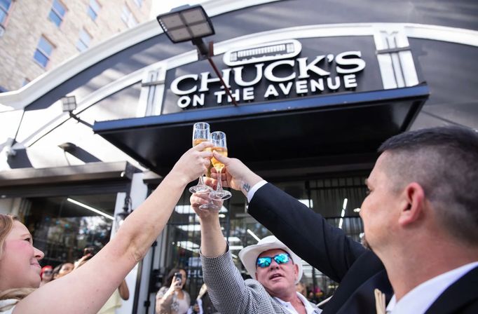 Four people clinking champagne flutes in a celebratory toast outside a black-and-white restaurant storefront on a busy city avenue, with onlookers and urban buildings in the background.