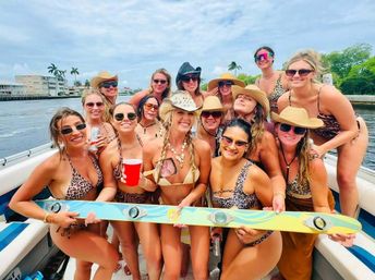 Group of women in swimsuits and sun hats smiling on a boat, holding a colorful wakeboard while cruising a palm-lined coastal canal