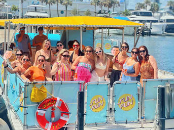 Smiling group of people on a colorful pontoon boat with a yellow canopy enjoying a sunny marina boat party, life ring and yachts in the background.