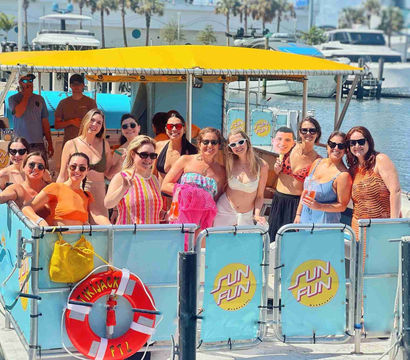 Smiling group of people on a colorful pontoon boat with a yellow canopy enjoying a sunny marina boat party, life ring and yachts in the background.