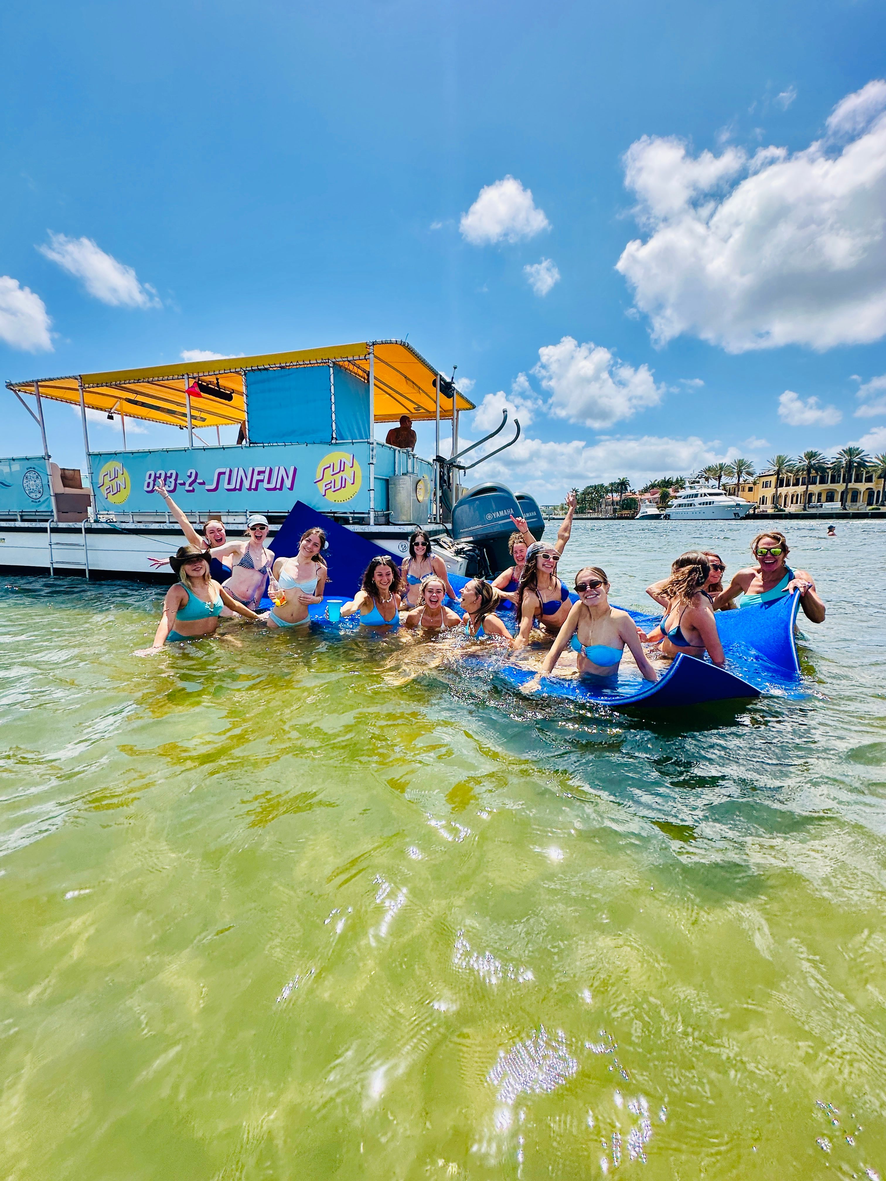 Boat party: group of people in swimsuits on a blue float in clear shallow green water beside a bright yellow‑canopy pontoon, palm‑lined waterfront and yachts under a sunny blue sky