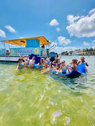 Boat party: group of people in swimsuits on a blue float in clear shallow green water beside a bright yellow‑canopy pontoon, palm‑lined waterfront and yachts under a sunny blue sky