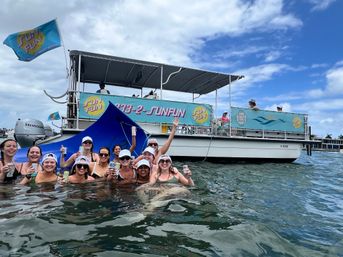 Group of friends in swimsuits cheering with drinks while floating in coastal Florida waters beside a colorful party pontoon boat with canopy and flags under a sunny, partly cloudy sky.