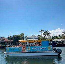 Colorful party pontoon boat with yellow canopy and passengers cruising a sunny tropical waterfront canal lined with palm trees and houses