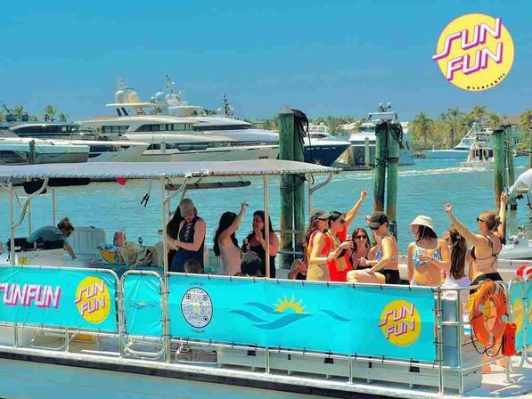 Group of people partying on a turquoise-decorated party boat at a sunny marina with luxury yachts, palm trees, and clear blue water in the background.