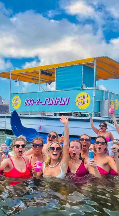 Cheerful group of women in colorful swimsuits waist-deep in a sunny coastal bay, holding canned drinks and cheering in front of a bright, decorated party pontoon boat with palm-lined shoreline
