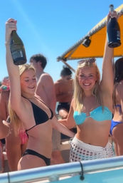 Two smiling women in bikinis holding up champagne bottles aboard a sunny boat party with a crowd and yellow canopy