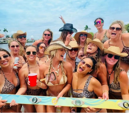 Cheerful group of women in swimsuits and cowboy hats partying on a boat, holding a red cup and colorful wakeboard with palm-tree waterfront in the background.