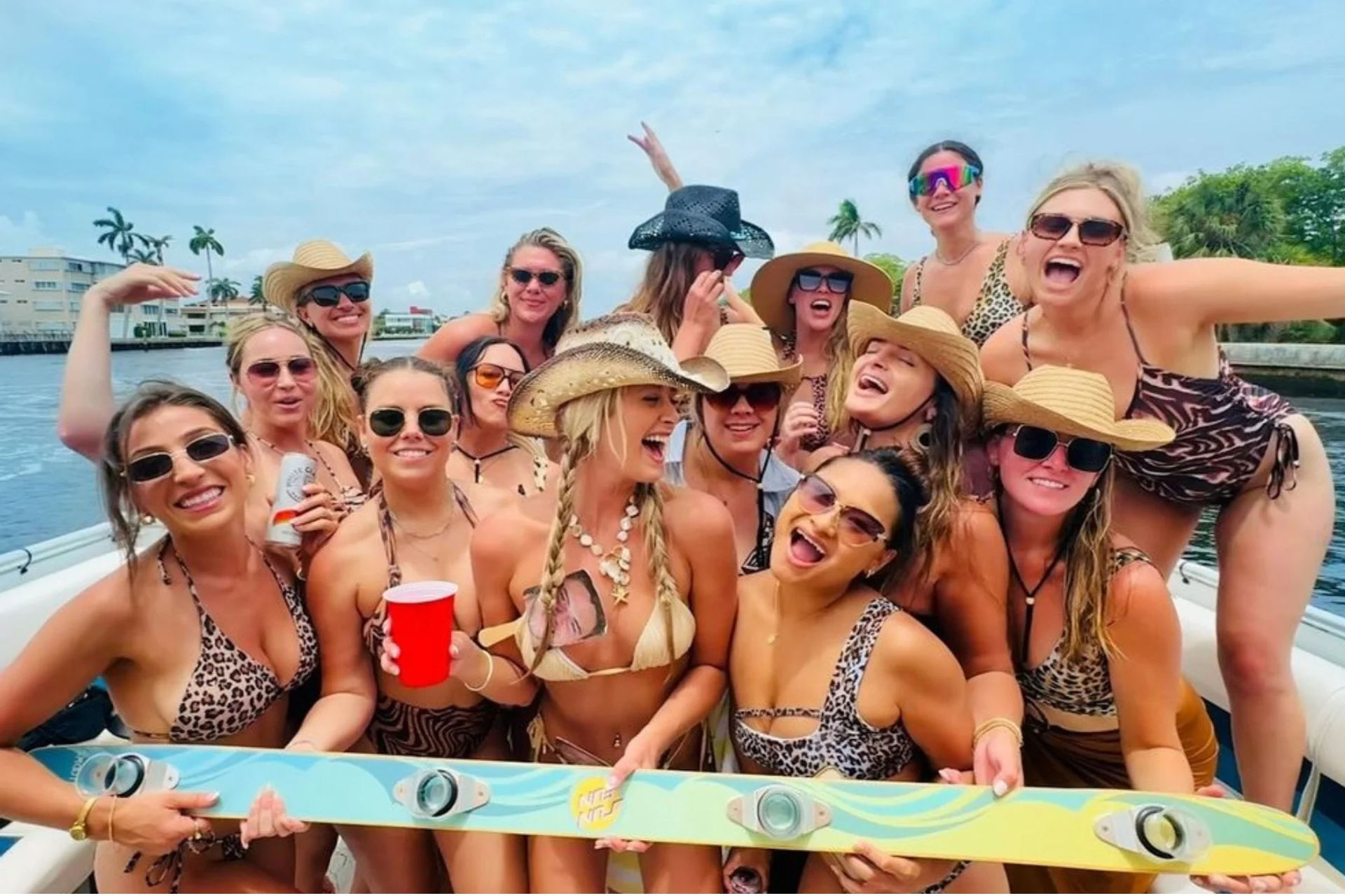 Cheerful group of women in swimsuits and cowboy hats partying on a boat, holding a red cup and colorful wakeboard with palm-tree waterfront in the background.