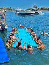Group of people partying around a bright blue floating bar in a marina, raising cups and cans while yachts glide by under a sunny sky.
