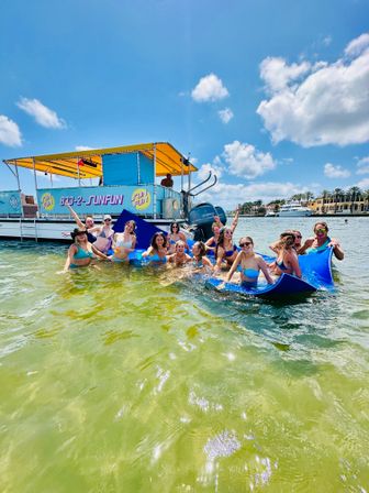 Smiling group in bikinis on a blue float in shallow green water beside a yellow‑canopied party pontoon boat, sunny sky and palm‑lined waterfront in background