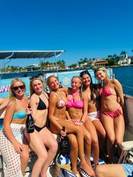 Six women in colorful bikinis posing with champagne bottles on a sunny boat party, palm-lined waterfront homes and clear blue sky in the background.