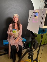 Smiling woman in a patterned headscarf seated in an indoor portrait studio, holding a colorful aura Polaroid while a camera on a tripod and a light projector cast neon patterns against a dark circular backdrop.
