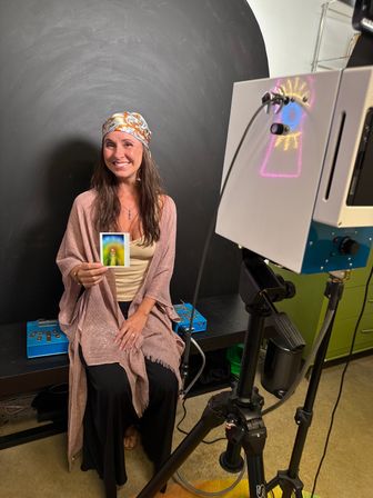Smiling woman in a patterned headscarf seated in an indoor portrait studio, holding a colorful aura Polaroid while a camera on a tripod and a light projector cast neon patterns against a dark circular backdrop.