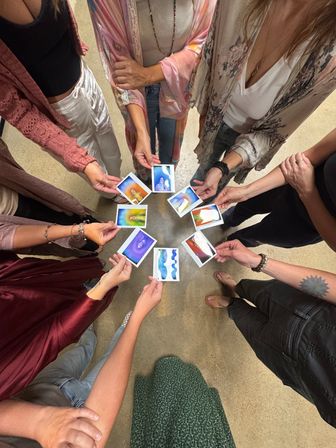 Top-down view of a circle of people in a wellness studio holding colorful aura-style Polaroid photos, boho clothing and hands extended toward the center.