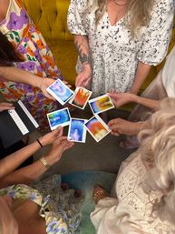 Top-down view of a cheerful circle of women indoors holding colorful Polaroid-style aura portraits, wearing floral dresses and jewelry near a mustard sofa.