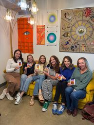 Six women seated on a mustard-yellow couch in a cozy wellness/art studio, smiling and holding colorful aura photos; boho wall tapestries, zodiac artwork and a crystal floor lamp add warm, eclectic decor.