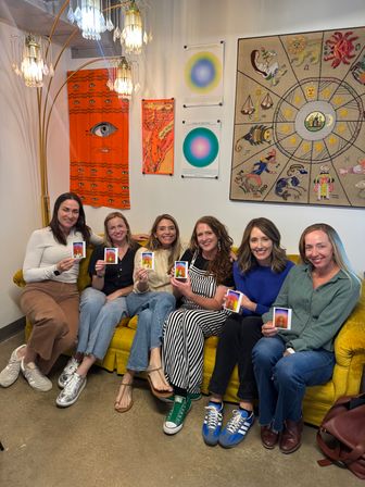 Six women seated on a mustard-yellow couch in a cozy wellness/art studio, smiling and holding colorful aura photos; boho wall tapestries, zodiac artwork and a crystal floor lamp add warm, eclectic decor.