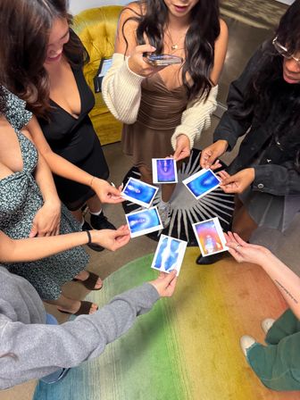 Friends at an indoor gathering compare blue-and-purple instant aura photos while standing on a rainbow rug near a mustard chair.