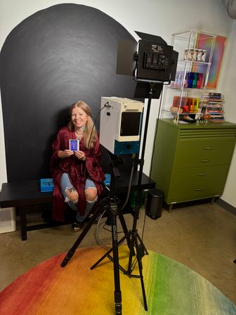 Indoor portrait studio scene: smiling person sits on a bench in front of a black arched backdrop holding an instant photo, with tripod-mounted lights, a rainbow circular rug and a green storage cabinet nearby.