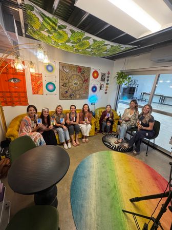 Eight women seated in a bright boho studio holding oracle cards on a yellow velvet couch and chairs, surrounded by zodiac wall art, an orange eye tapestry, a neon eye sign, hanging plant, and a rainbow rug.