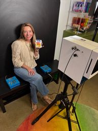 Smiling woman in a casual blouse and jeans sits in a small indoor portrait studio against a black backdrop, holding a colorful instant photo as a camera on a tripod and studio lights capture the shot, with shelves of supplies and a rainbow rug adding bright accents.