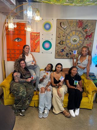 Six women seated on a mustard-yellow couch in a cozy art studio, smiling and holding colorful aura photos; background shows a zodiac tapestry, orange eye poster, circular color charts and hanging crystal lamps.
