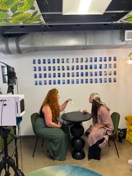 Two women seated across a black sculptural table in a bright creative studio, one showing a colorful card while a grid of blue-tinted portrait prints lines the wall, exposed metal ductwork and floral ceiling panels overhead, with studio lighting and camera gear nearby.