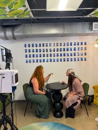 Two women seated across a black sculptural table in a bright creative studio, one showing a colorful card while a grid of blue-tinted portrait prints lines the wall, exposed metal ductwork and floral ceiling panels overhead, with studio lighting and camera gear nearby.