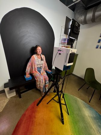 Woman in a colorful kimono seated with eyes closed in an indoor photo-studio portrait setup—black arched backdrop, rainbow gradient rug, and camera with lighting on a tripod in the foreground.