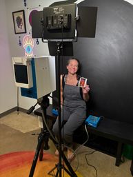 Smiling woman in overalls sits in an indoor portrait studio against a black backdrop, holding an instant photo print while tripod-mounted camera, studio lights, neon eye wall art, and framed prints surround her.