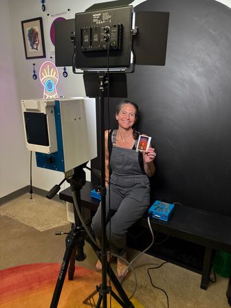 Smiling woman in overalls sits in an indoor portrait studio against a black backdrop, holding an instant photo print while tripod-mounted camera, studio lights, neon eye wall art, and framed prints surround her.