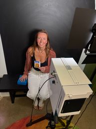 Beaming woman seated on a bench in a small portrait studio holding a tiny colorful instant photo, with a black backdrop, studio lighting and camera equipment visible