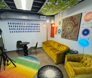 Eclectic studio lounge interior with mustard-yellow velvet sofa and armchair, rainbow round rug, black sculptural table with green chairs, zodiac wall art, hanging lamps and neon blue eye sign.