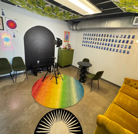 Vibrant indoor photo studio with tripod and softbox, rainbow round rug, neon eye sign, polaroid wall, green dresser and mustard sofa