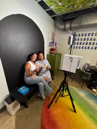 Two friends smile and show instant photos while seated against a black arched backdrop in a colorful photo studio — tripod-mounted camera and light, rainbow rug, shelving with products and a wall of small prints behind them.