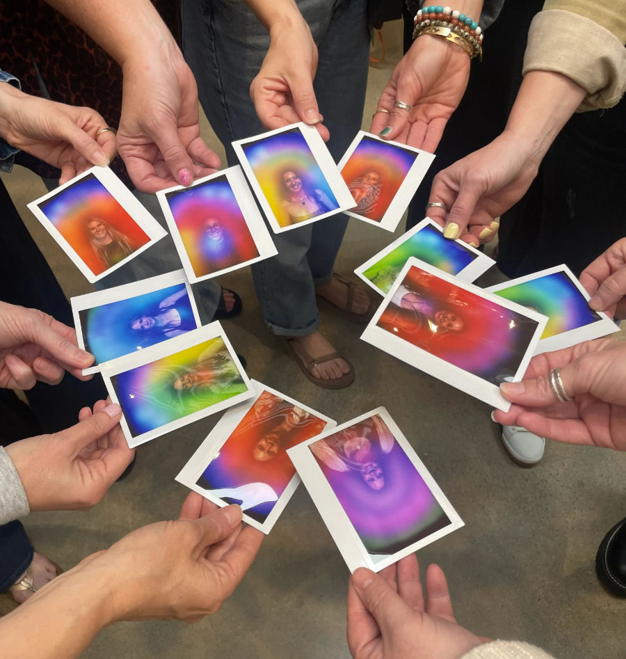 Overhead shot of a group holding instant Polaroid-style photos in a circle, each portrait surrounded by vibrant rainbow aura colors, hands and feet visible on a concrete floor — colorful group keepsake moment.